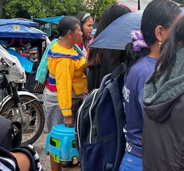 Indigenous woman in traditional yellow clothing waiting in line at an outdoor market in the rain.