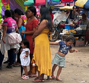 Indigenous women and children walking through a crowded outdoor marketplace with colorful stalls.