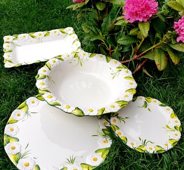 Hand-painted ceramic dinnerware set with 3D daisy flowers displayed on green grass by pink rhododendrons.
