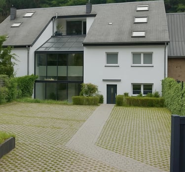 Maison moderne blanche avec grande véranda en verre, toit gris et allée pavée entourée de verdure.