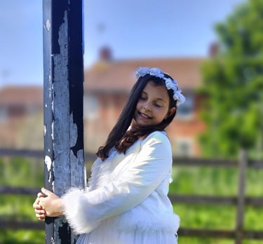 Close-up of a young girl with long brown hair, wearing a white dress and a floral white headband, smiling outdoors on a sunny