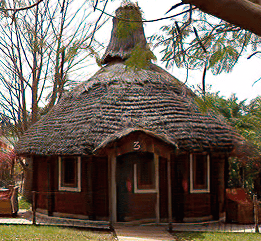 Traditional round African hut with a thatched roof and brown walls in a lush tropical garden.