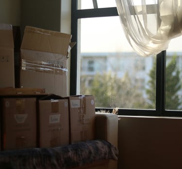 Stacked cardboard moving boxes in a bright room next to a window with white curtains.