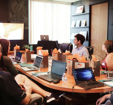 Colleagues attending a corporate business meeting in a modern office boardroom with laptops and a presenter.
