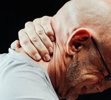 Bald man grimacing in pain while holding his stiff neck to relieve muscle tension and soreness.