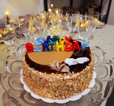Chocolate birthday cake with nut topping and colorful Happy Birthday candles on a glass stand.