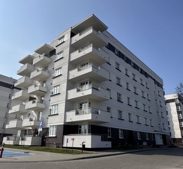 Nowoczesny budynek mieszkalny z jasną elewacją i szarą podstawą, z parkingiem z przodu.