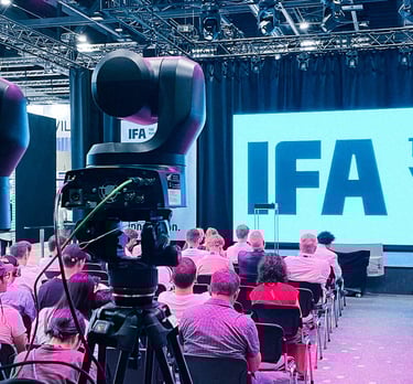 Publikum in Konferenzraum vor großem IFA-Bildschirm, Kamera auf Stativ filmt Veranstaltung.