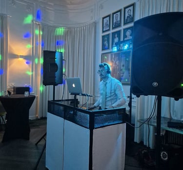 Professional event DJ with laptop and large speakers performing at a party venue with disco lights.