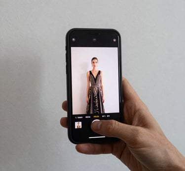 Una mano sosteniendo un smartphone que muestra una galer&iacute;a de im&aacute;genes de alta costura, con un fondo de pared blanca y sombras suaves.