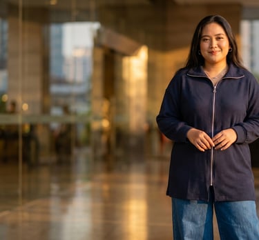A smiling woman wearing a navy blue zip-up jacket and jeans standing in a modern office lobby at sunset.