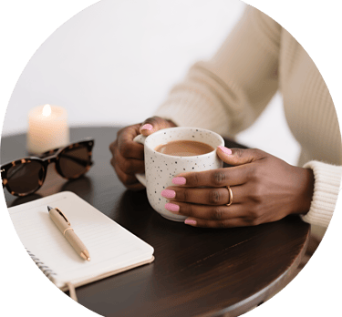 A woman holds a ceramic coffee mug on a table with a notebook, pen, and sunglasses.