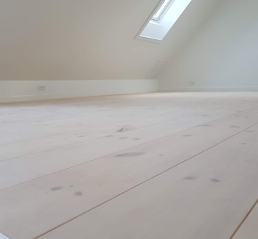 Tomt loftsrum med lyst fyrretræsgulv og et moderne ovenlysvindue.