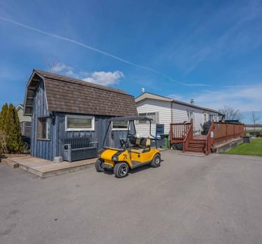 Yellow golf cart parked outside a blue shed and mobile home with wooden deck under a blue sky.