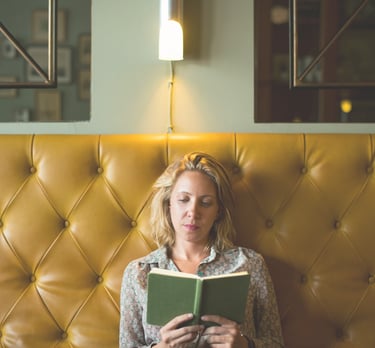 A blonde woman reading a book while sitting against a yellow tufted leather banquette in a cafe.