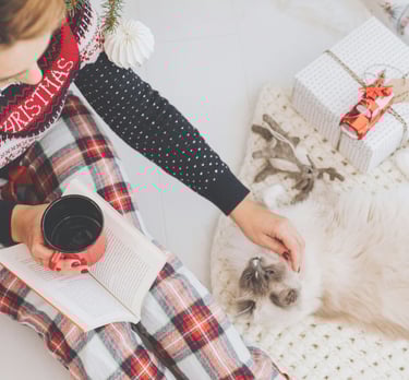 Moteris su kalėdine pižama ir megztiniu sėdi ant grindų, glosto katę, rankoje puodelis.