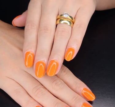 Professional manicure featuring vibrant orange gel nail polish and gold rings on a woman's hands.