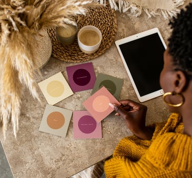 A woman holds colorful affirmation cards on a desk with a digital tablet and coffee.