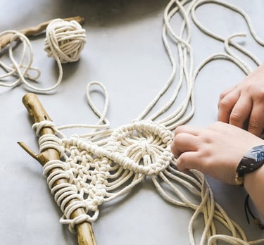 Manos elaborando un tapiz de macramé con cordón de algodón blanco sobre una rama de madera natural.