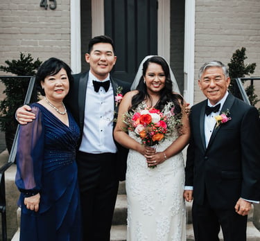 Smiling bride and groom posing with parents in formal wedding attire outside a brick house.