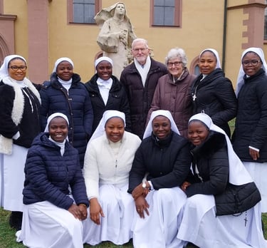 A group of Catholic nuns in white habits and black jackets posing with a senior couple in front of a church.