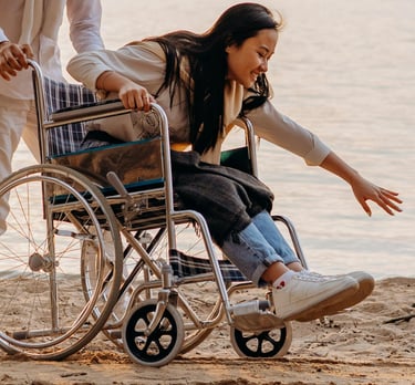 Donna in sedia a rotelle sulla spiaggia, accompagnata, mentre tocca l’acqua del mare