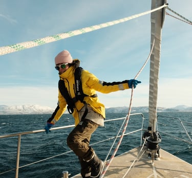 Maria på seilbåtdekk i Lofoten, gul jakke og sikkerhetsvest, vinterfjell i horisonten.