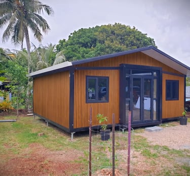 Modern wood-paneled tiny home or prefab cabin with black trim in a lush tropical garden setting.