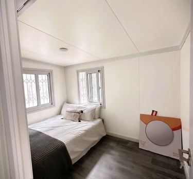 A minimalist tiny house bedroom with a white bed, dark wood floors, and dual windows.
