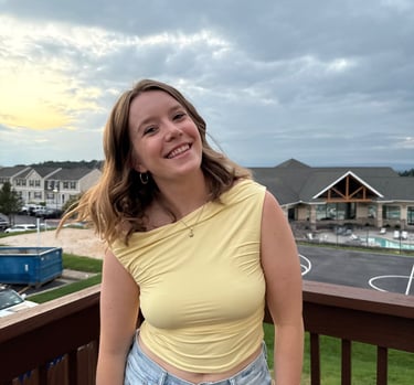 Smiling woman in a yellow top and denim jeans posing on a wooden balcony at sunset.