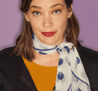 A smiling woman with brown hair and a white and blue polka dot scarf, wearing a mustard shirt and black cardigan, against a