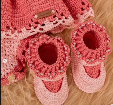Handmade pink crochet baby booties and dress with pearl bead accents on a soft faux fur background.