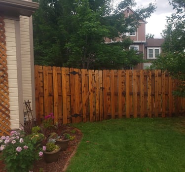A wooden cedar privacy fence with a gate stands in a backyard garden with green grass and pink flowers.