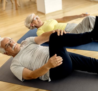 Active senior couple performing knee-to-chest stretches on yoga mats in a bright fitness studio.