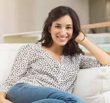 A smiling woman in a patterned blouse and jeans sits comfortably on a white sofa.