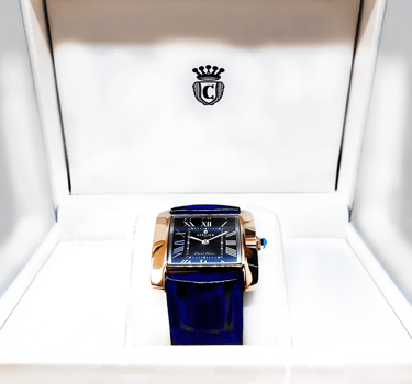 Luxury rectangular rose gold watch with a blue leather strap and Roman numeral dial in a white display box.
