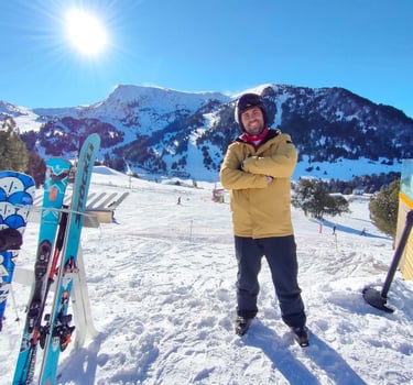 Smiling male skier in yellow jacket standing on a snowy mountain slope with blue skis under a bright sun.