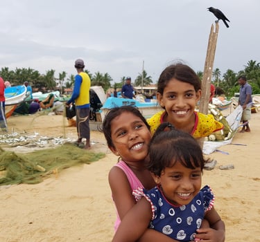 Three smiling Sri Lankan girls playing on a sandy beach near fishing boats and nets in a coastal village.