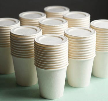 A professional product photograph of neat stacks of biodegradable paper soup cups with secure lids, standing on a clean mint green surface. The lighting is soft and natural, emphasizing the pristine quality and eco-friendly textures of the material.