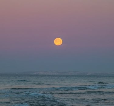 a full moon rising over the ocean with a pink sky