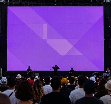 Perspective shot of a large, sophisticated crowd at a music festival in a Spanish / Latin American city, silhouettes against a massive LED screen displaying minimalist geometric patterns in vibrant electric violet. Atmosphere of experience and high-level production.