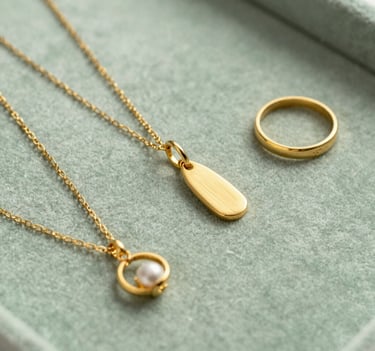Photography of delicate gold jewelry items arranged on a soft sage green velvet surface. The composition is artistic and editorial, reflecting a North American / Western European luxury brand aesthetic. Bright, airy lighting captures the warm glow of the gold.