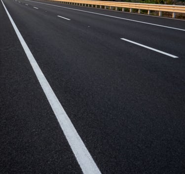 A wide-angle professional photograph of a newly constructed six-lane highway in India. The image features crisp, bright white thermoplastic road markings and reflective studs stretching towards the horizon. The lighting is the golden hour of sunset, highlighting the smooth black asphalt. South Asian landscape in the background.
