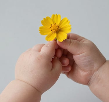 A close-up photography of a baby's tiny hands holding a single yellow flower. The background is a clean, solid light lead grey surface. The focus is sharp on the texture of the skin, reflecting high-quality professional photography.