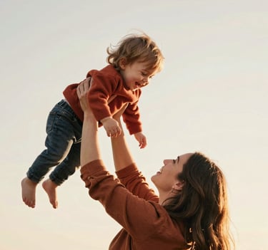 A medium shot of a mother lifting her toddler in the air, both beaming with joy. The scene is bathed in sun-drenched warmth, with the child wearing a terracotta-colored sweater against a soft off-white sky.