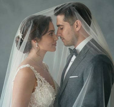 Medium shot of a bride and groom sharing a quiet, intimate moment behind a sheer cream white veil. Soft focus, romantic lighting, slate grey-blue color tones, sophisticated and emotional, Hispanic / Spanish-speaking couple.