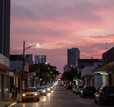 An artistic wide shot of an urban street in Maracay, Venezuela at dusk, captured with a cinematic lens, featuring blurred city lights in salmon pink and rose against a dark sky.