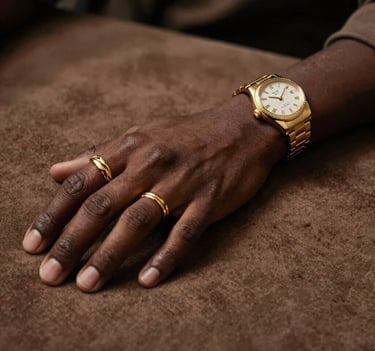 A close-up, high-fashion shot of P.E.P YLS's hands adorned with intricate gold rings and a luxury watch, resting on a textured #8D765D brown fabric. The lighting is cinematic and focused, highlighting the artistic detail and the contrast between the dark skin tones and the golden accessories.