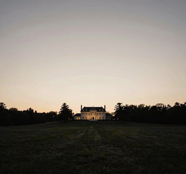 A wide-angle landscape photograph of a North American / European countryside estate at dusk. The sky is a gradient of warm grey and off-white. In the distance, the silhouettes of the manor are dark charcoal. The overall feel is one of timeless elegance and minimalist luxury.