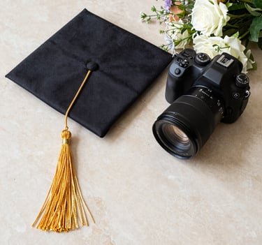 A creative flat lay photography of a graduation mortarboard with a luxurious golden tassel next to a professional DSLR camera and a bouquet of fresh flowers, set on a Light Beige marble surface.